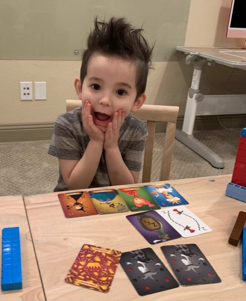 Child with his hands over his cheeks making a playful shocked expression. Cards from the game Sleeping Queens are on the table in front of him.