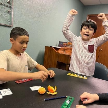 Two children are playing one of the games for math intervention, Odd Todd's Even. One child has both arms in the air and an excited expression. The other is looking down at his dice.